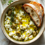 Shallow bowl filled with an olive oil-based dip featuring crumbled feta, fresh herbs, chopped olives, and lemon zest, garnished with herbs and served alongside slices of toasted bread.